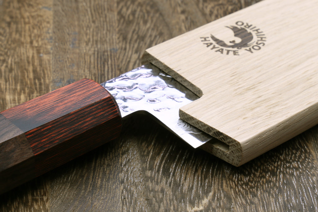 Santoku knife blade partially inserted into oak wood saya cover on light wood