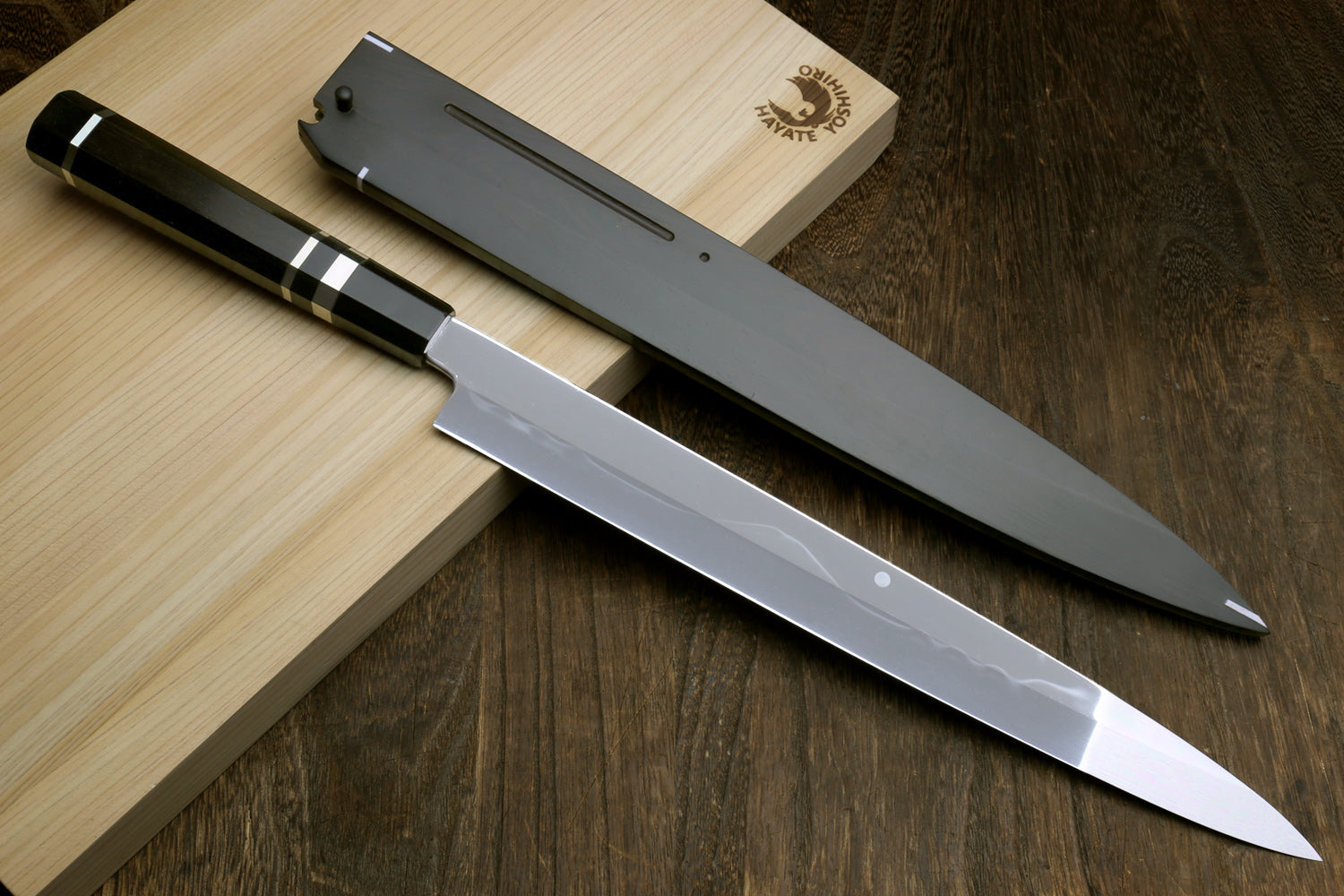 Japanese chef’s knife with mirror blade and ebony handle, shown with black scabbard.