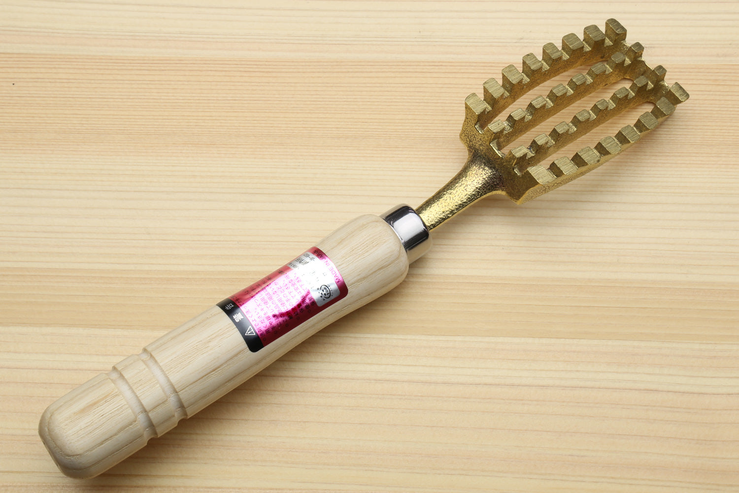 Fish scaler tool with light wood handle and gold metal teeth, pink label on handle