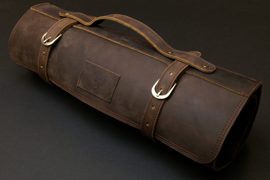 Brown leather knife roll case with top carry handle, side label patch, and metal rings.