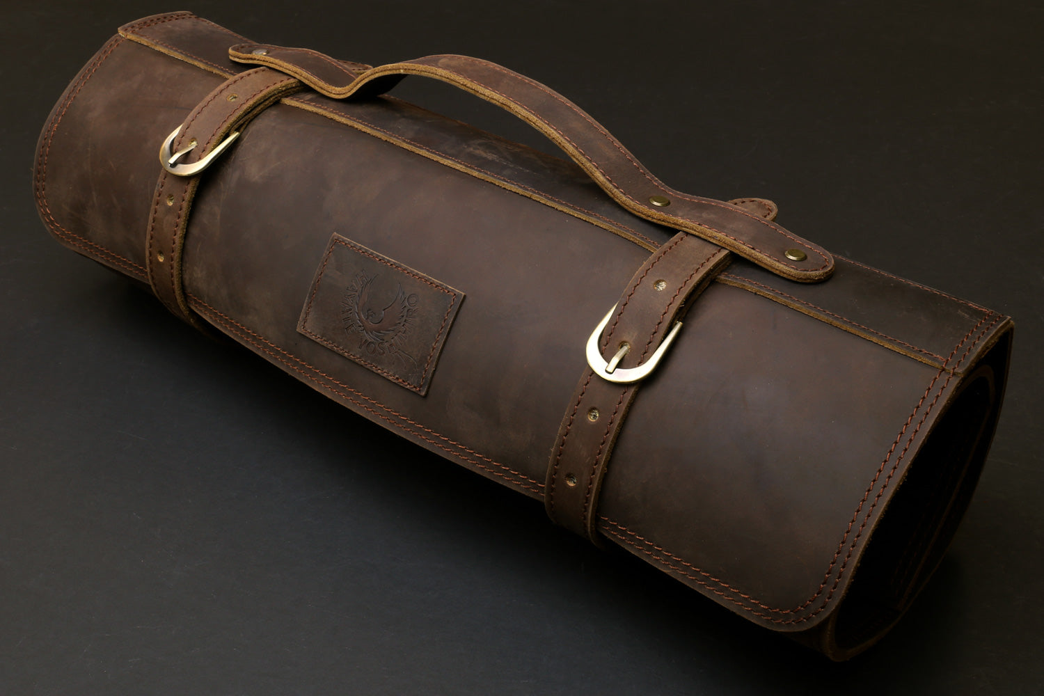 Brown leather knife roll case with top carry handle, side label patch, and metal rings.