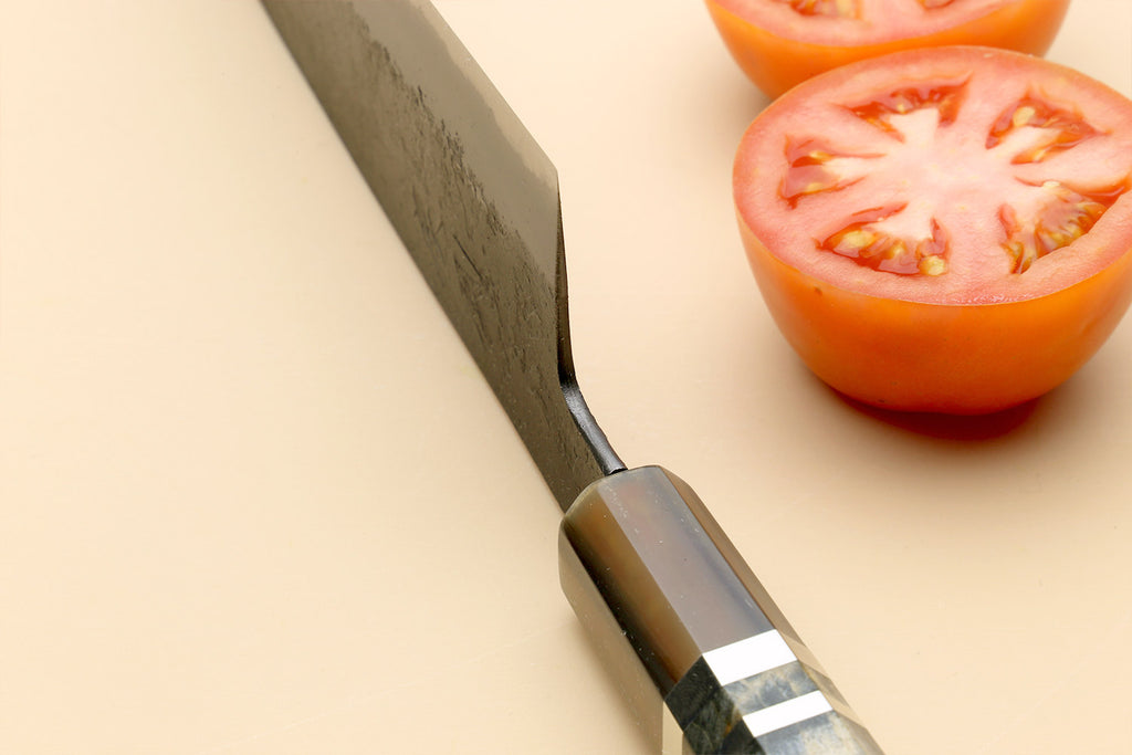 Santoku knife close-up on blue-steel blade and dark handle, by tomato halves