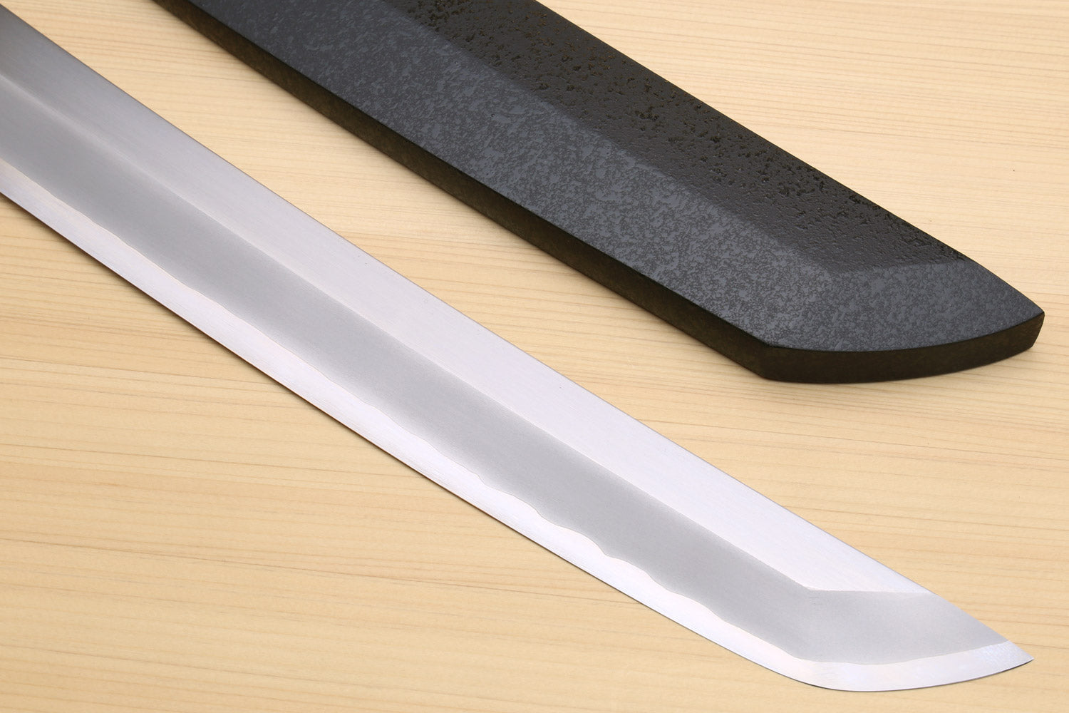 Tuna knife blade and black saya, long silver edge on a light wood surface