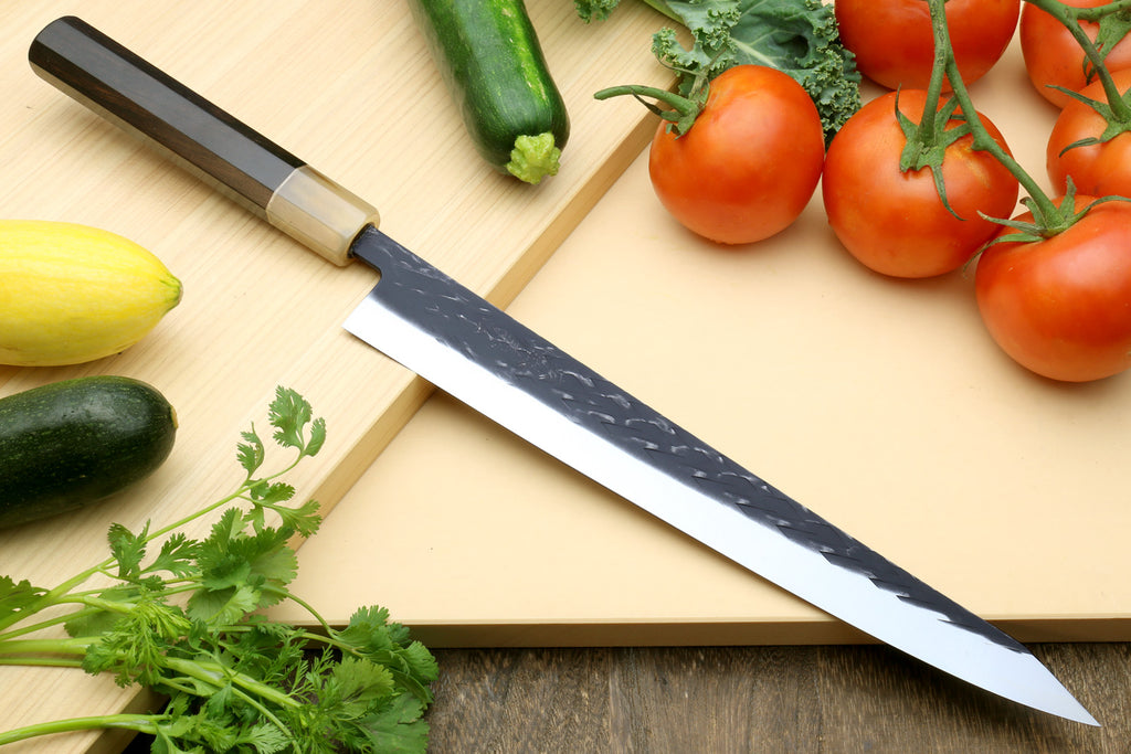 Stainless chef knife with speckled blade and dark handle, with tomatoes, zucchini, and herbs