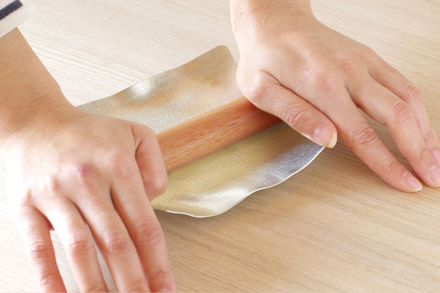 Hands pressing a flexible tin plate around a wooden rolling stick