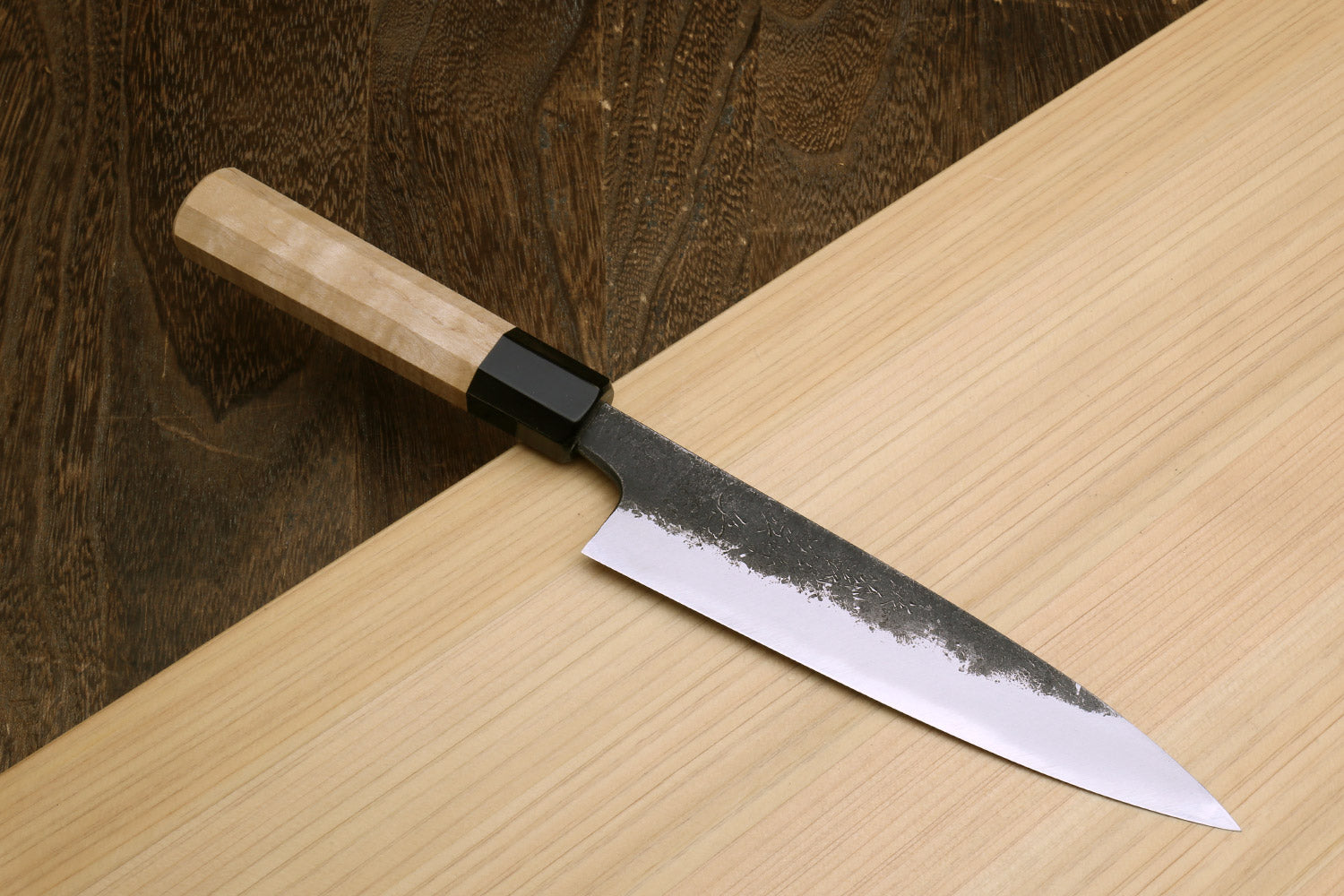 Petty utility knife with dark speckled blade and black bolster on light wood handle