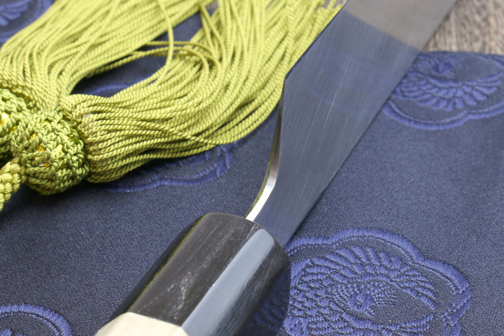 Japanese chef knife blade close-up with white edge and magnolia handle, green tassel cord.
