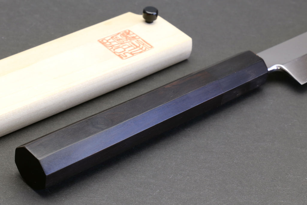 Sushi chef knife with mirror blade and dark ebony handle, resting on light saya.
