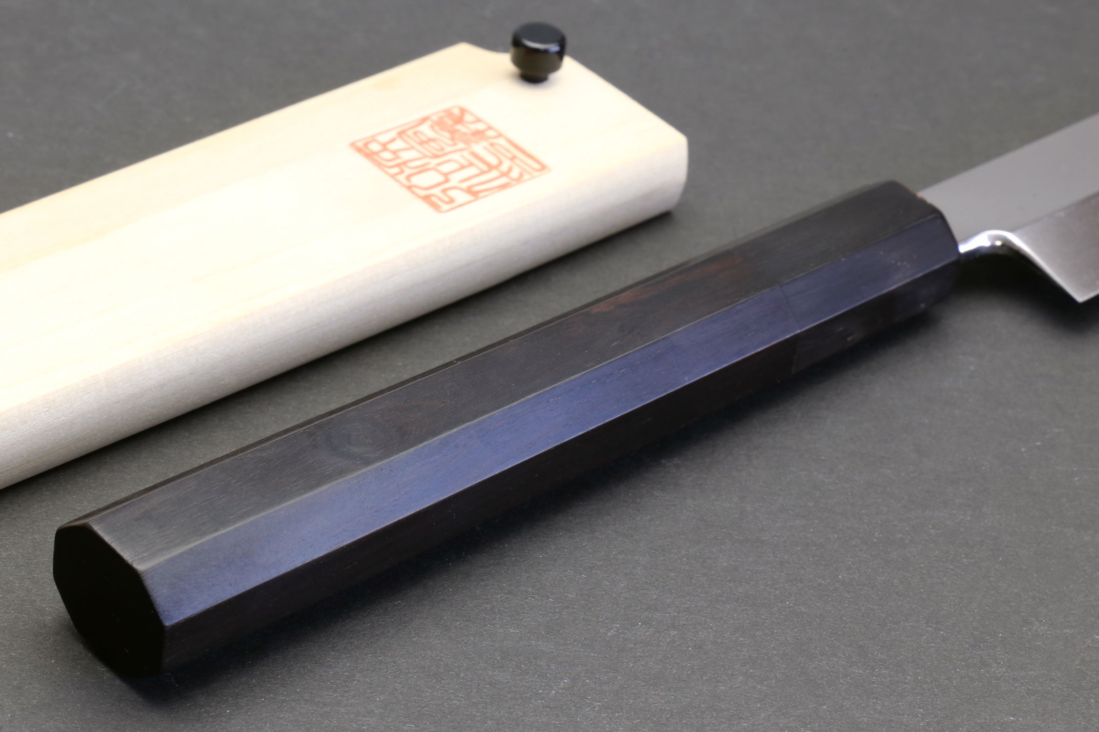 Sushi chef knife with mirror blade and dark ebony handle, resting on light saya.