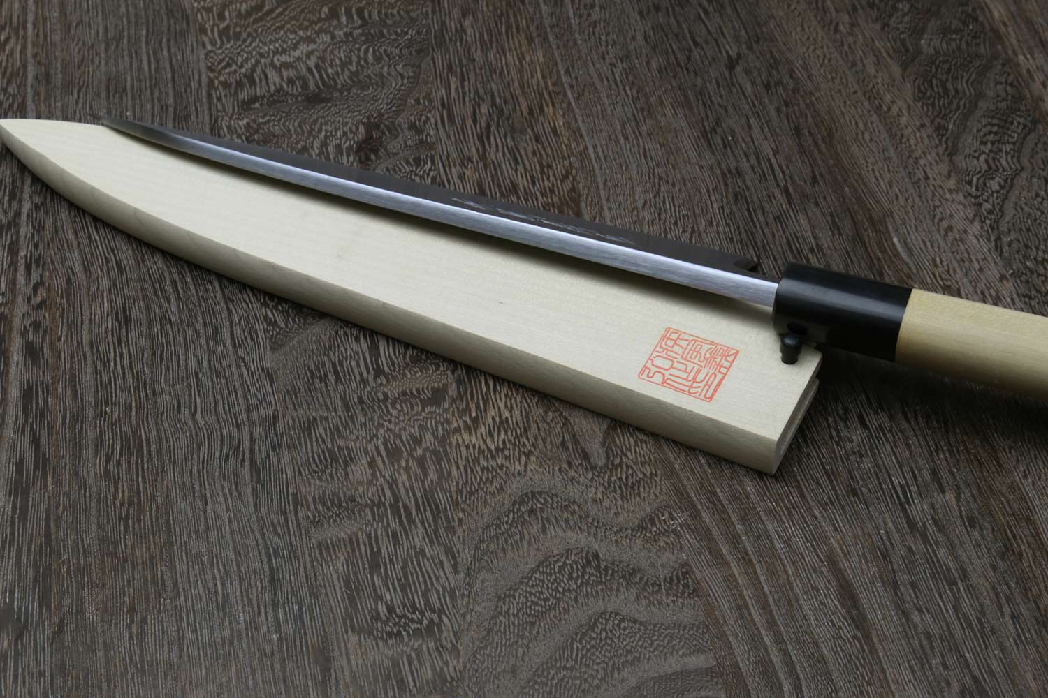 Sashimi filet knife with silver blade resting in light wood saya, black bolster
