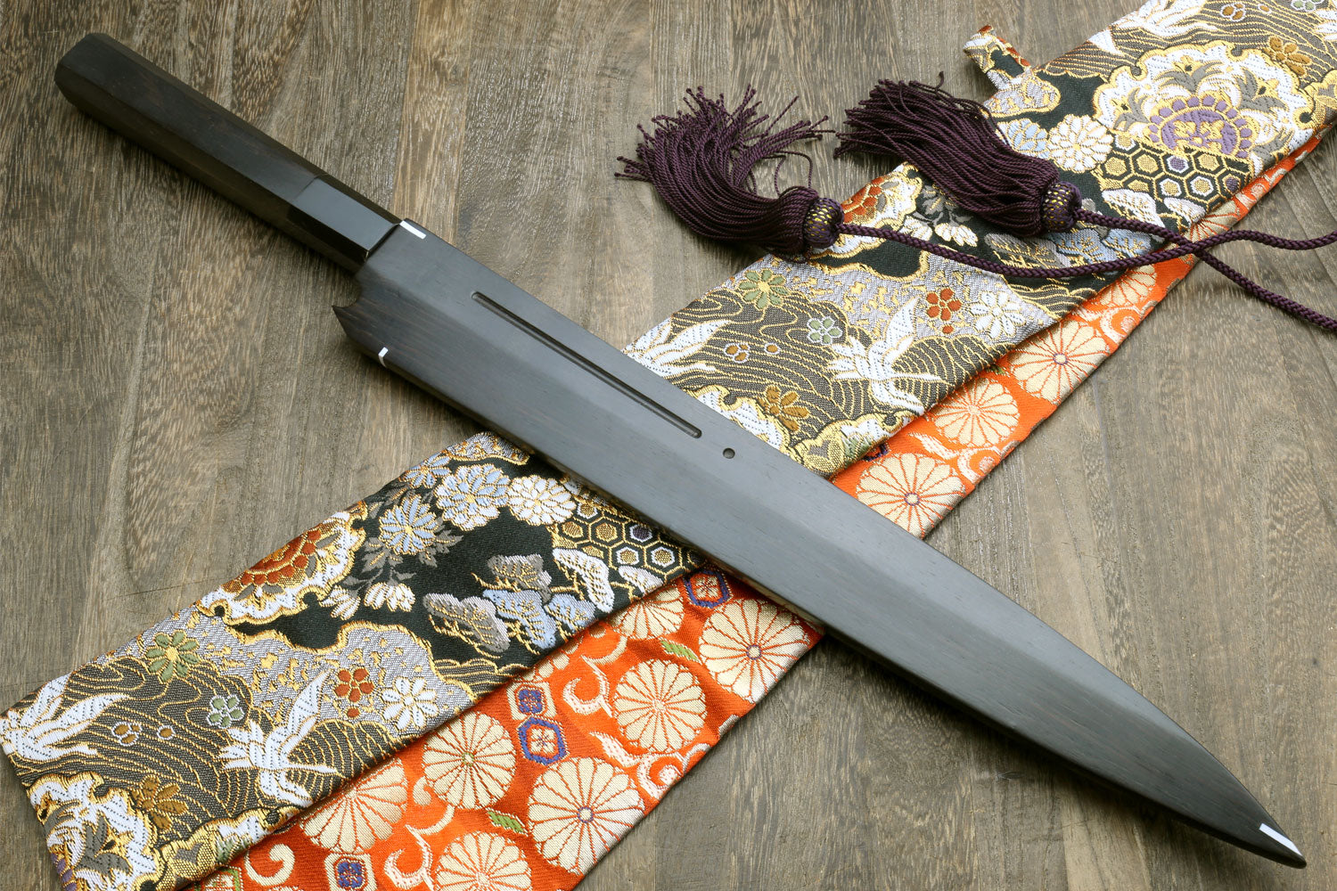 Sashimi knife and saya laid out on patterned fabric, dark handle and purple cord tassel