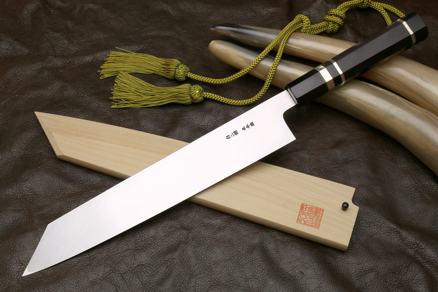 Japanese-style kitchen knife with wavy steel blade, black grip, green cord tassel, saya