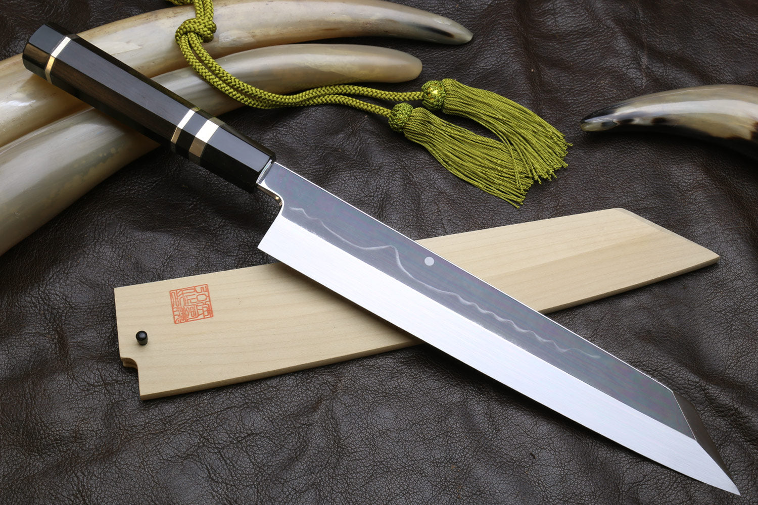 Japanese-style kitchen knife set with wavy steel blade, light wood sheath, and green cord tassel