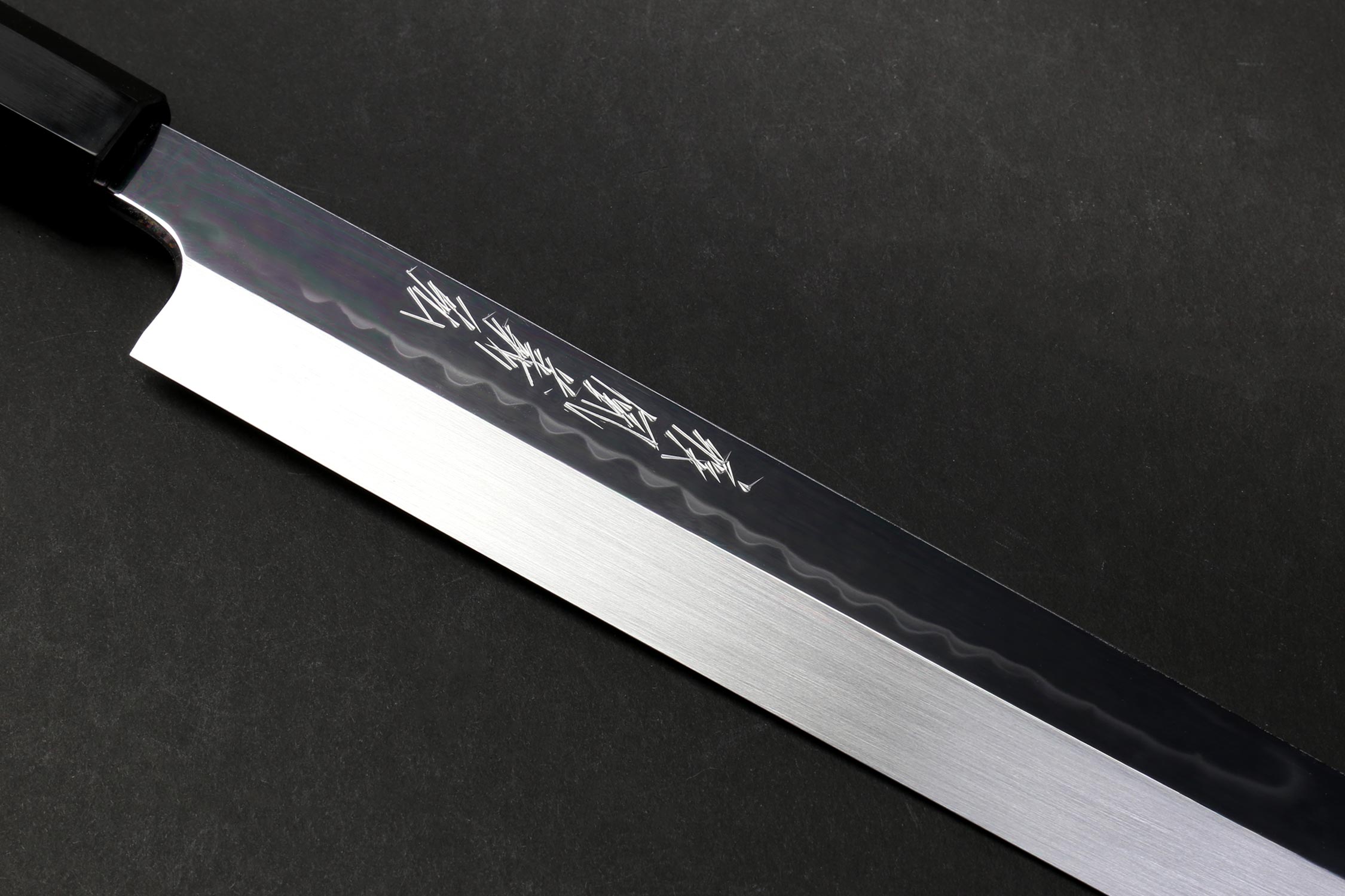 Japanese knife blade close-up with mirror-finished steel and etched pattern near the spine.