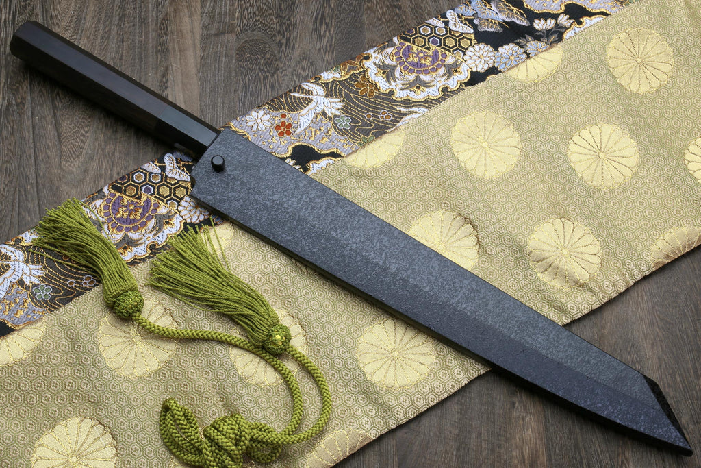 Japanese knife with silver mirrored blade and dark handle on patterned cloth with green tassels.