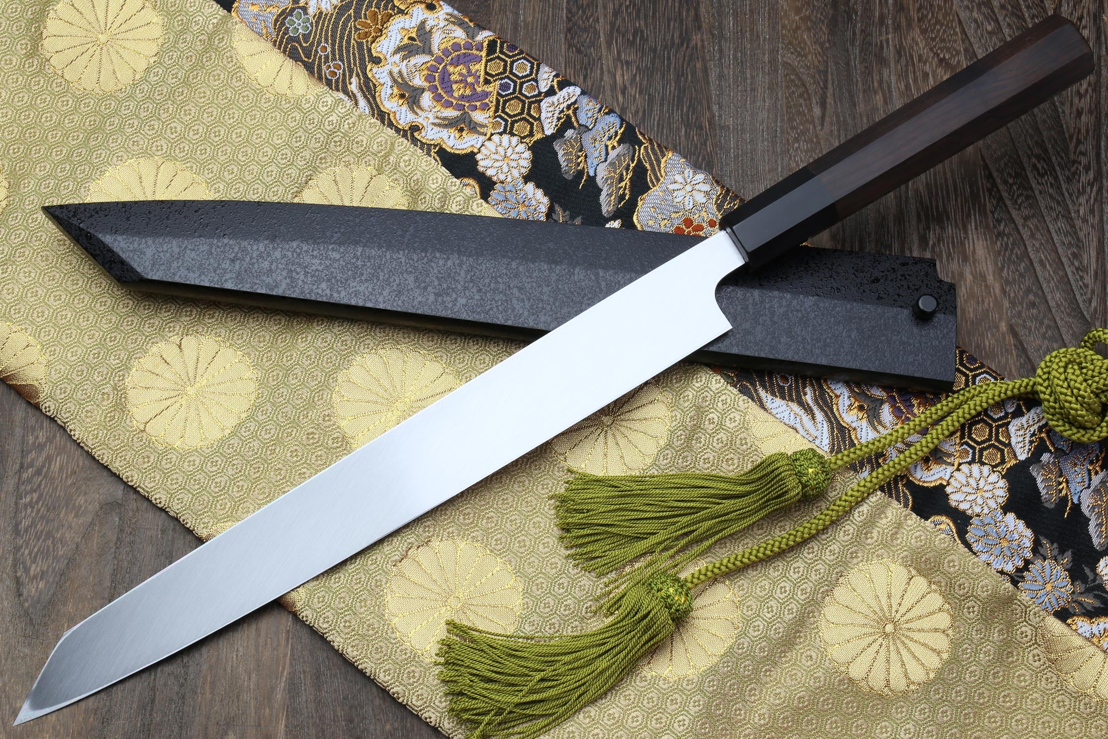 Japanese knife with silver mirrored blade and dark handle, shown crossed over cloth with tassels.