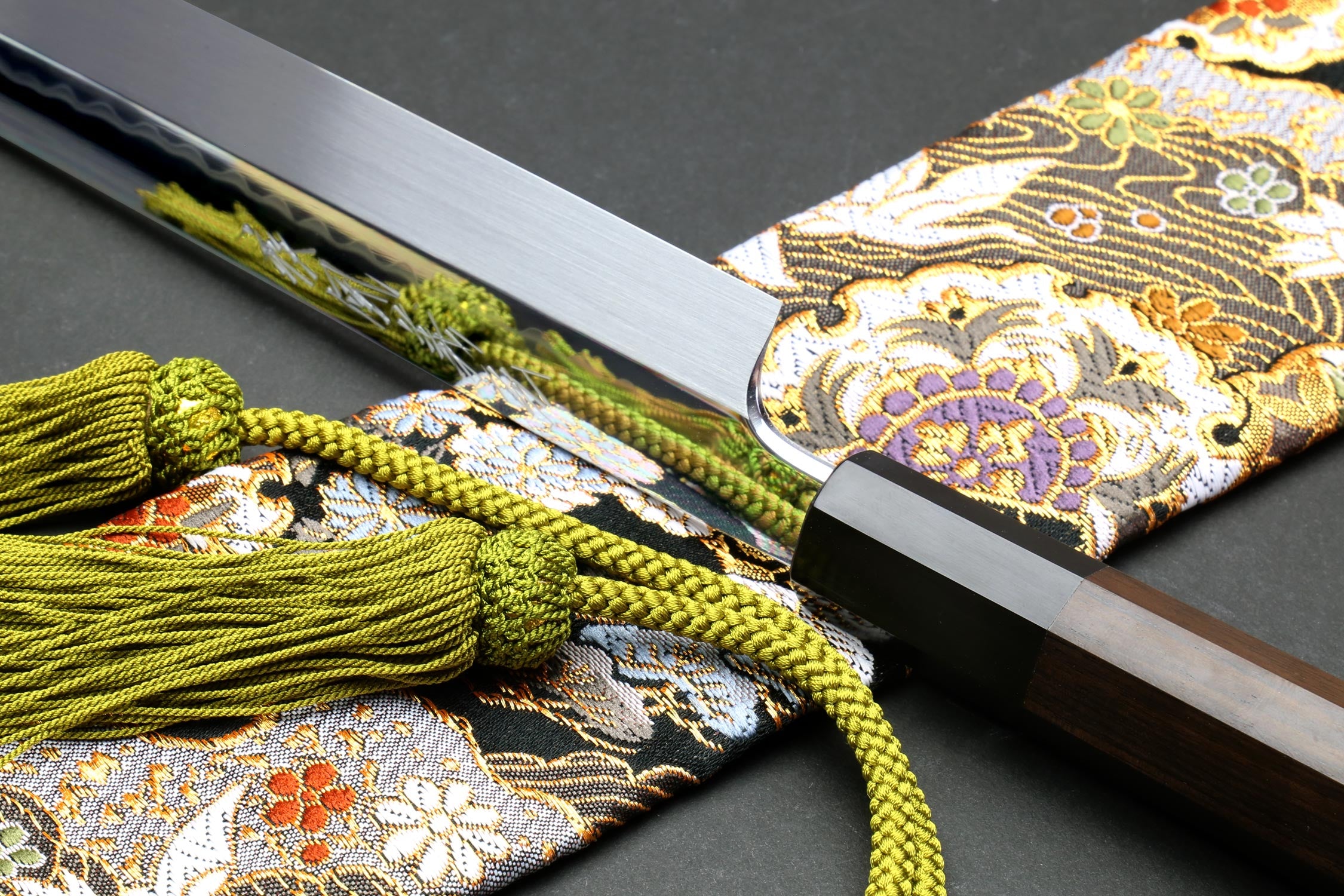 Japanese knife with silver blade and black handle beside green tassel cord and patterned cloth.