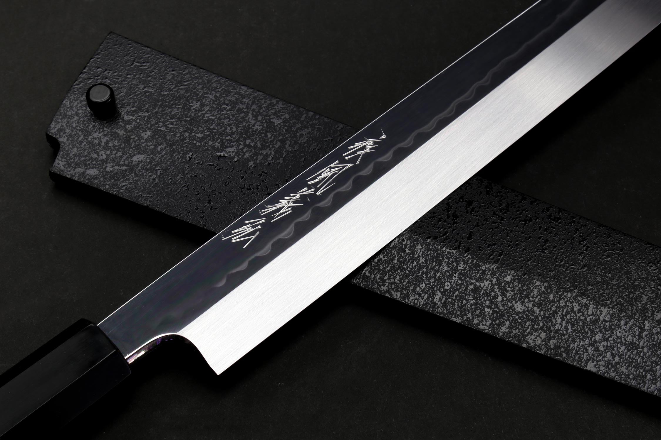 Japanese knife with mirror-finished blade and dark handle, close-up on engraved marking near spine.