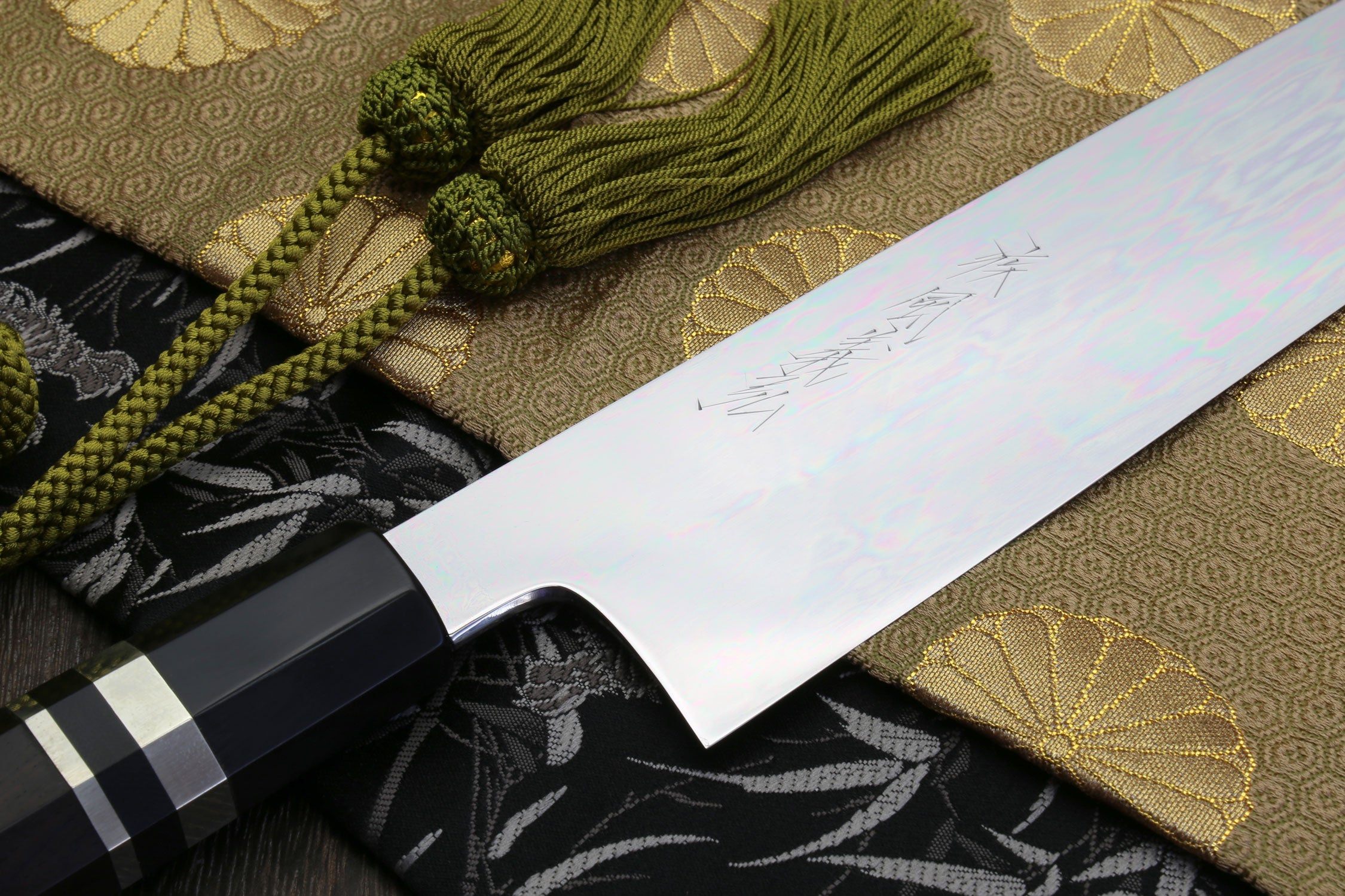 Gyuto chef knife detail with mirror-polished blade, ebony handle, and green cord tassels