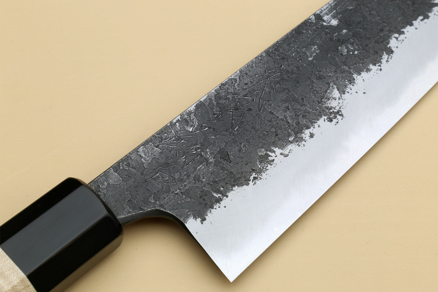 Close-up of white steel gyuto blade with nashiji finish and dark bolster, no handle.