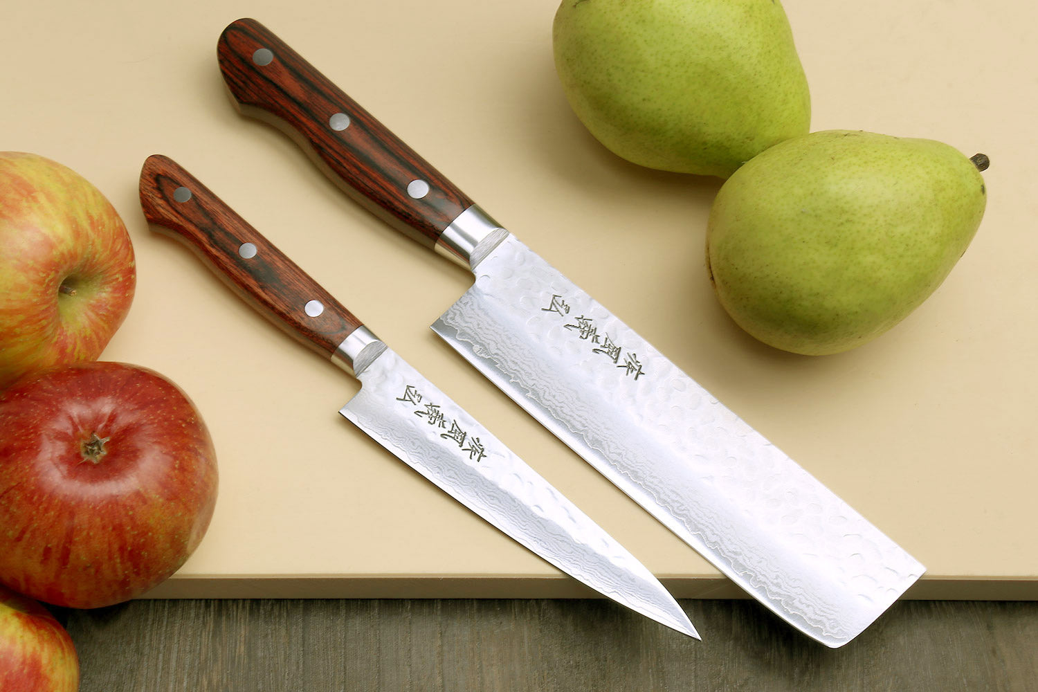 Nakiri and petty knives with dark wood handles and hammered Damascus blades, on board.